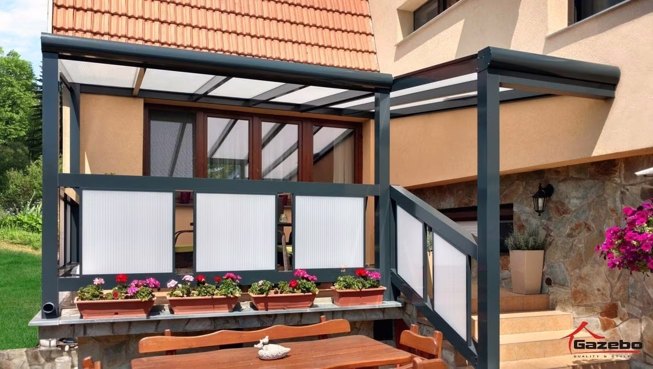 Anthracite Economic Class aluminium pergola with glazed sides beside a stone house with flowers
