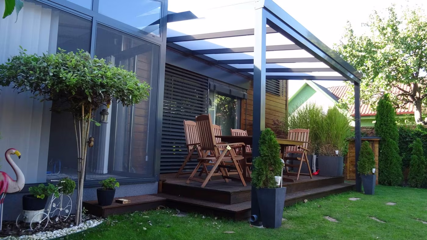 Anthracite Economic Class aluminium pergola over a wood deck with a dining set by a hedge