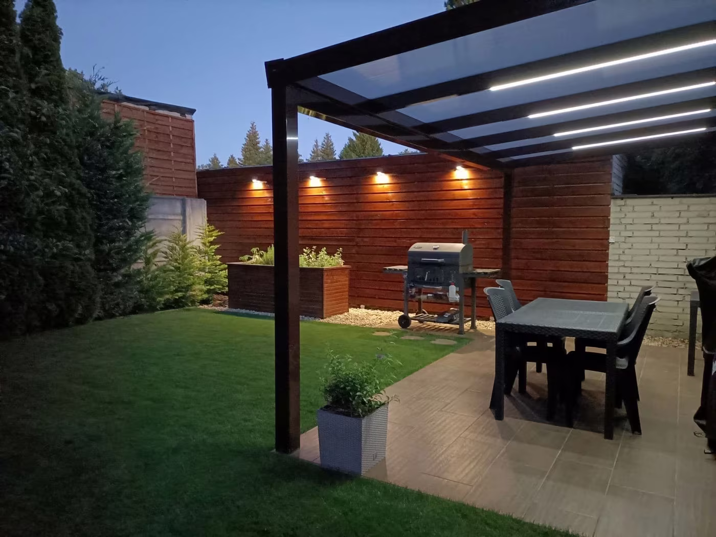 Illuminated anthracite Economic Class aluminium pergola over a paved terrace with a grill in the evening