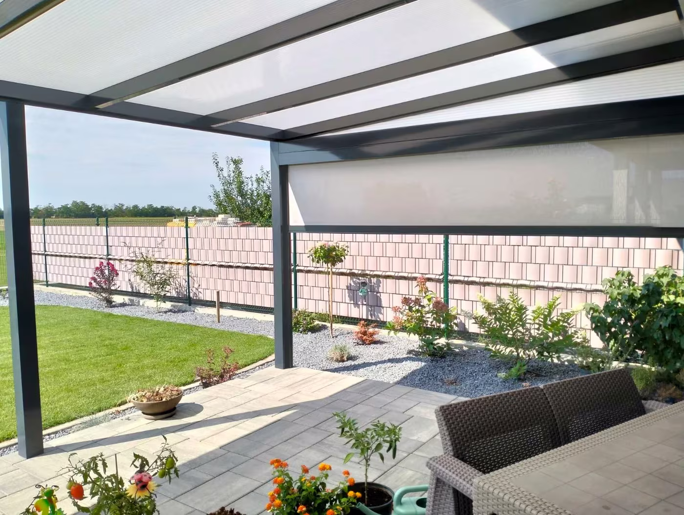 Anthracite Economic Class aluminium pergola along a fence with rattan seating and flowers
