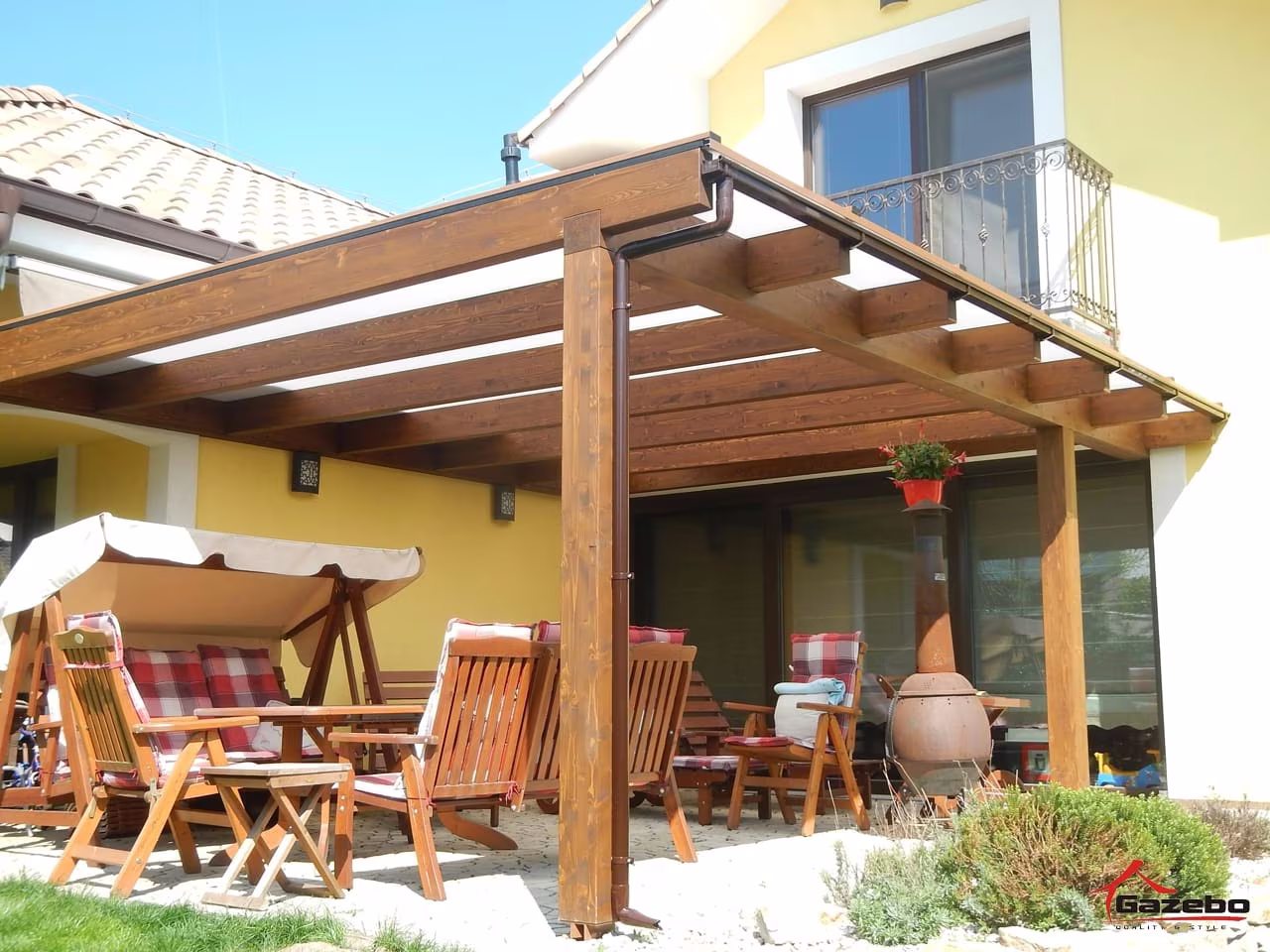 Fenix wooden pergola over a terrace with garden furniture and a swing seat by a yellow house